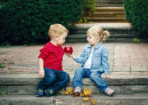 2 kids on a side walk sharing a red apple.